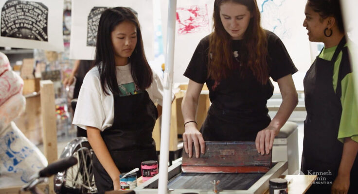 Two people watch as a person pulls ink on a screen at a printing table.