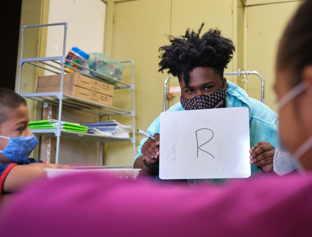 A dark skinned person wearing a face mask and holding up a whiteboard with the letter R written on it to two students