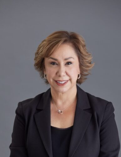 A light skinned Latina with short brown hair wearing a black blazer and black top with silver earrings and necklace