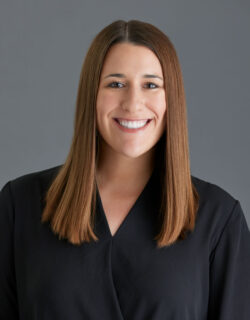 Headshot of Devon Williams, a white woman with medium length, straight brown hair. She’s smiling at the camera and wearing a V-neck black blouse.