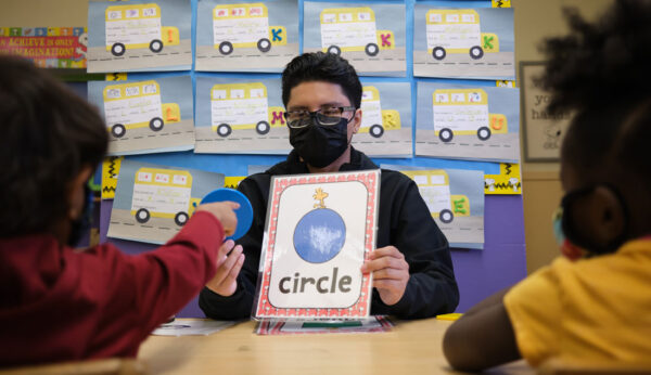 Edward, Villanueva, and Early Literacy King, wearing a black face mask, holding up a sign that reads “circle” with a picture of a blue circle in one hand. He holds a plastic blue circle in another hand that a child points to.