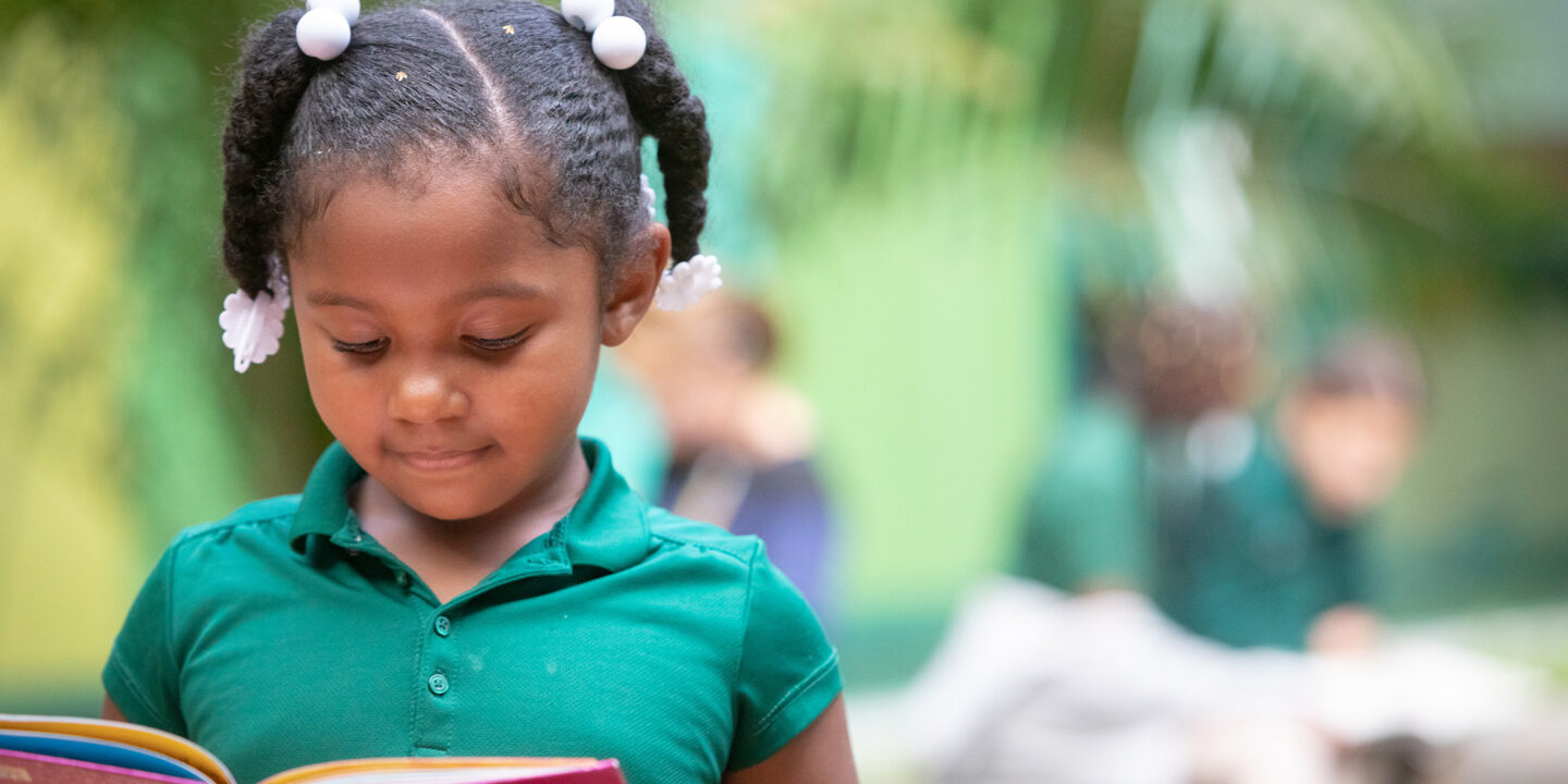 A young Black girl smiling while she looks down at an open book, reading.