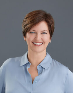 Headshot of Jen Rainin, a white woman with wavy brown hair. She’s smiling at the camera, wearing a black blouse and a pendant necklace.
