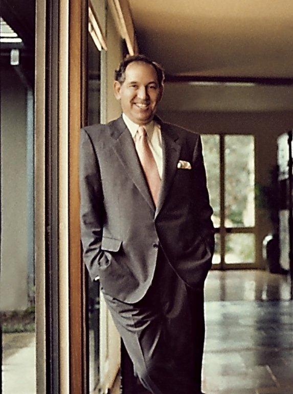Kenneth Rainin wearing a gray suit with a peach tie, leaning against a door jam and smiling at the camera.