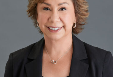 A light skinned middle-aged woman with short brown hair wearing a black blazer, black top, and silver earrings and necklace.