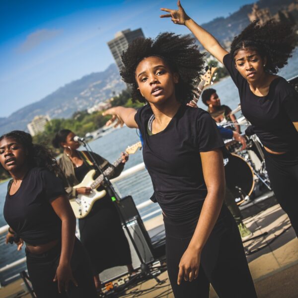 Three young Black female dancers performing on Lake Merritt in front of a band including a keyboardist, guitarist and drummer.
