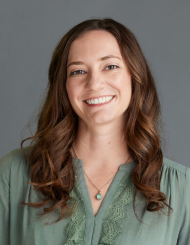 Headshot of Megan De Trane, a white woman with long, wavy brown hair. She’s smiling at the camera, wearing a light green blouse with a matching pendant necklace.