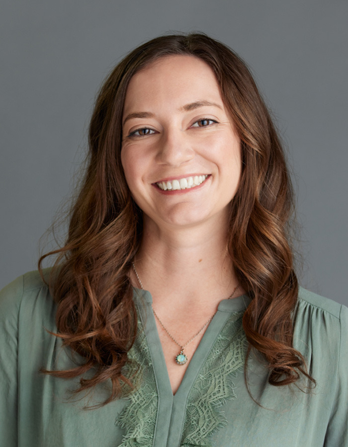 Headshot of Megan De Trane, a white woman with long, wavy brown hair. She’s smiling at the camera, wearing a light green blouse with a matching pendant necklace.