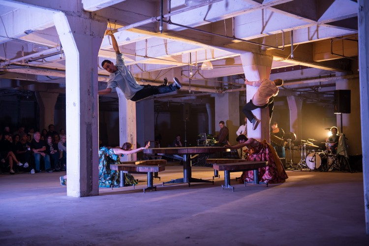 In what looks like a commercial garage, a site-specific dance performance is taking place between two large support beams. Two female dancers in teal and magenta dresses are on opposite sides of an outdoor table with benches, bent over and pushing into the table forcefully. One male dancer is climbing a support beam. Another male dancer is holding onto the second support beam with both legs raised in the air.