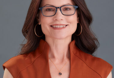Headshot of Shelley Trott, a white woman with long, wavy auburn hair. She’s smiling at the camera, wearing a burnt orange dress, hoop earrings and glasses.