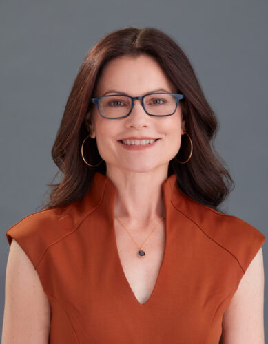 Headshot of Shelley Trott, a white woman with long, wavy auburn hair. She’s smiling at the camera, wearing a burnt orange dress, hoop earrings and glasses.