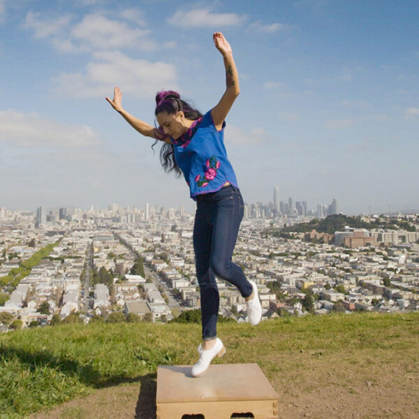 A Chicana-Native dancer standing on the tiptoes of one foot, with arms spread wide above her head. In the background is dense city housing and downtown San Francisco high-rise buildings.