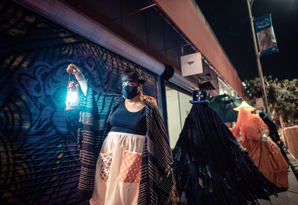 A Black woman wearing a face mask and holding a lantern, leading a procession in the dark.
