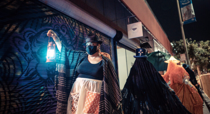 A Black woman wearing a face mask and holding a lantern, leading a procession in the dark.
