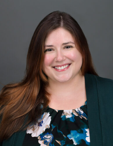 Headshot of Victoria Pozzato, a white woman with long, brown hair. She’s smiling at the camera, wearing a green and blue floral blouse under a dark green cardigan.