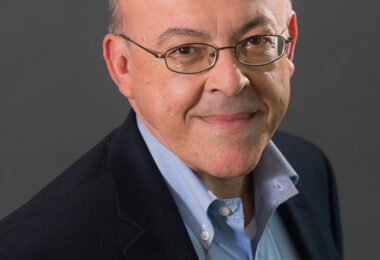 Headshot of Jerry Taillon, a mature white male with glasses. He’s smiling at the camera wearing a light blue shirt and a dark blue jacket.