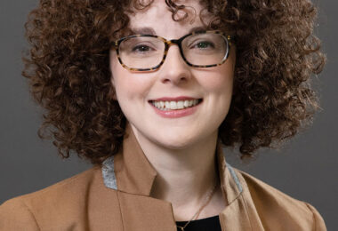 Headshot of Karina Ryan, a white woman with curly brown hair. She’s smiling at the camera, wearing Tortoiseshell glasses, a black blouse, a camel blazer and a pendant necklace.