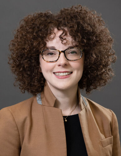 Headshot of Karina Ryan, a white woman with curly brown hair. She’s smiling at the camera, wearing Tortoiseshell glasses, a black blouse, a camel blazer and a pendant necklace.