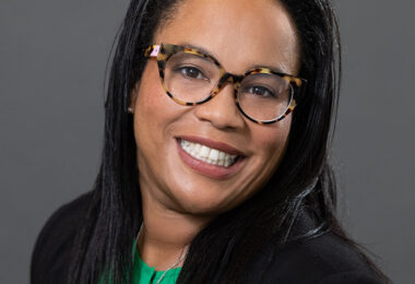 Headshot of Miyesha Perry, a black woman with long, straight black hair. She's smiling at the camera, wearing a green blouse, black blazer and houndstooth framed glasses.