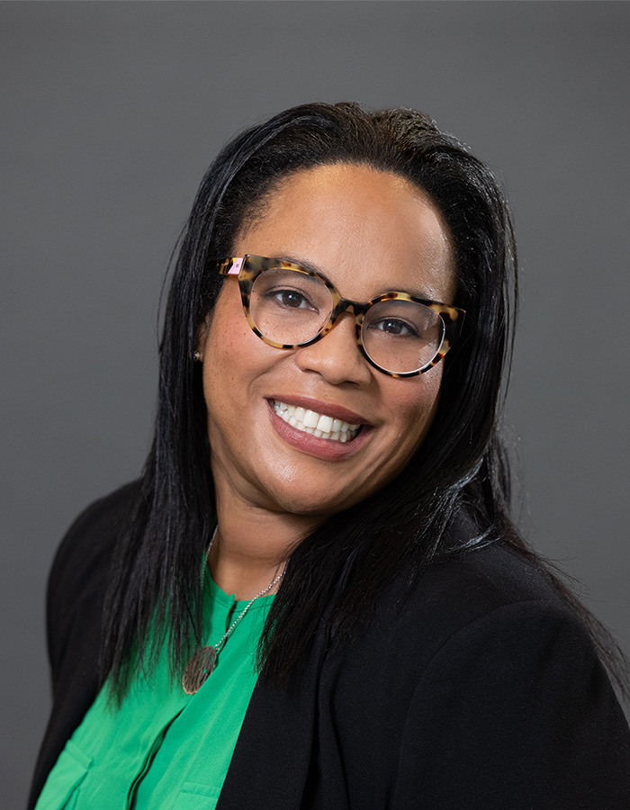 Headshot of Miyesha Perry, a black woman with long, straight black hair. She's smiling at the camera, wearing a green blouse, black blazer and houndstooth framed glasses.