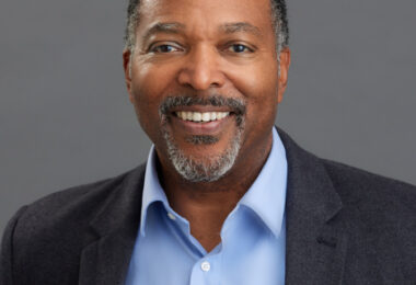 Headshot of Ted Russell, an African-American male with medium-toned brown skin and closely cropped salt and pepper gray hair and goatee. Appearing before a gray background, he’s wearing a blue-colored oxford shirt underneath a dark grey blazer.