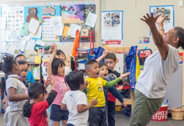 A teacher and young students stand in a classroom and sing joyfully and extend their arms upward.