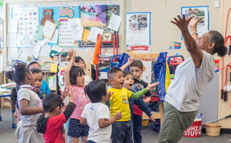 A teacher and young students stand in a classroom and sing joyfully and extend their arms upward.