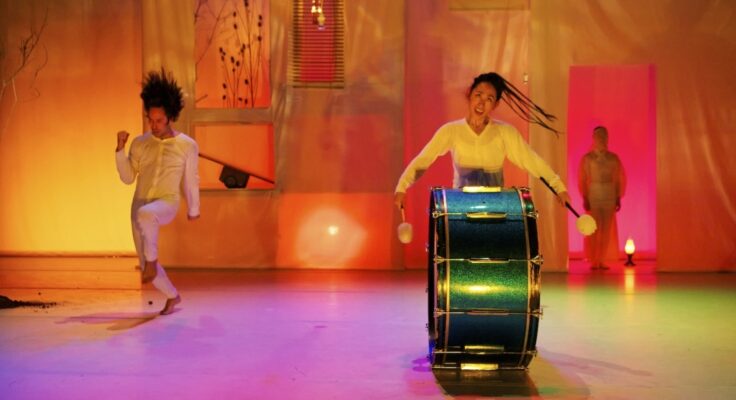 A colorful stage in shades of yellow, pink and purple. A woman beats a very large drum in the foreground as a man dances in the background.