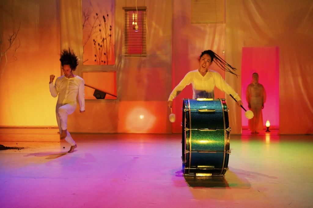 A colorful stage in shades of yellow, pink and purple. A woman beats a very large drum in the foreground as a man dances in the background.