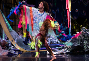Dancer mid-movement on a stage wearing a garment that is silver with fluorescent trim. In the background colorful fabrics hang and drape the stage.