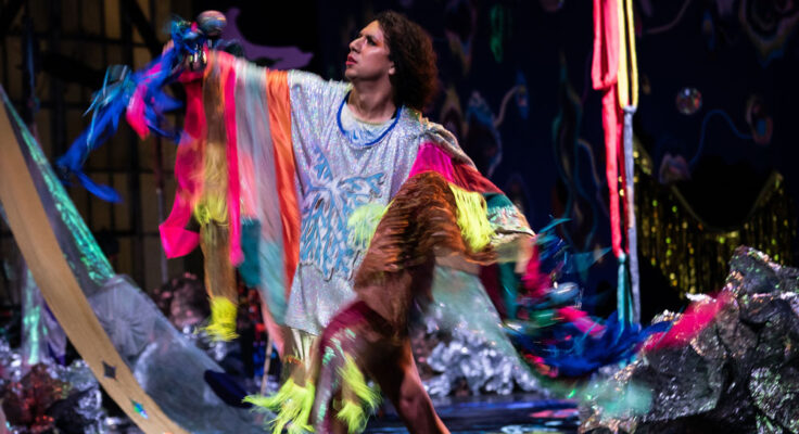 Dancer mid-movement on a stage wearing a garment that is silver with fluorescent trim. In the background colorful fabrics hang and drape the stage.