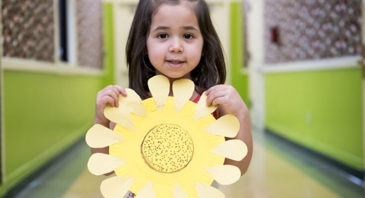 A young girl smiling and holding up a large sunflower she made out of colored paper