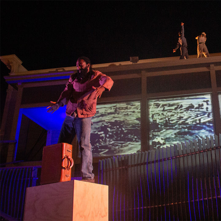 A site specific dance in Bayview-Hunter’s Point where three dancers are on top of a roof and one dancer is dancing on a wooden block with a briefcase.