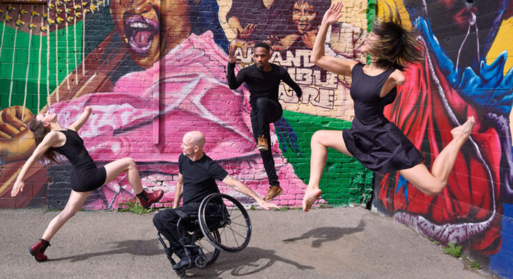Four disabled artists dressed in black are in various states of motion; two on the ground and two jumping in the air in front of a colorful mural.