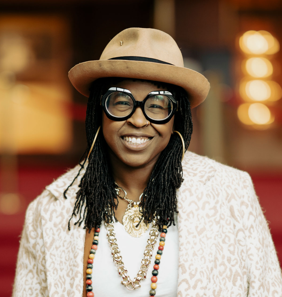 a queer Black interdisciplinary artist, wearing a short rimmed beige hat and large eyeglasses.