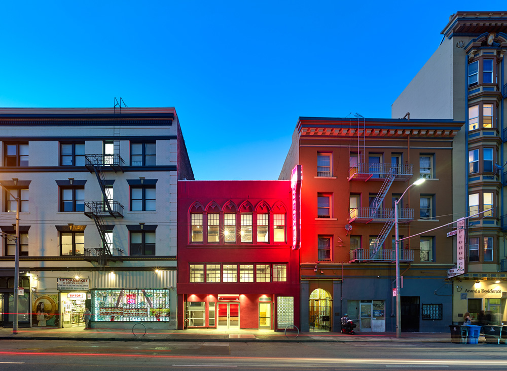A three story magenta colored building with light shining from all of its windows sandwiched between other buildings in San Francisco.