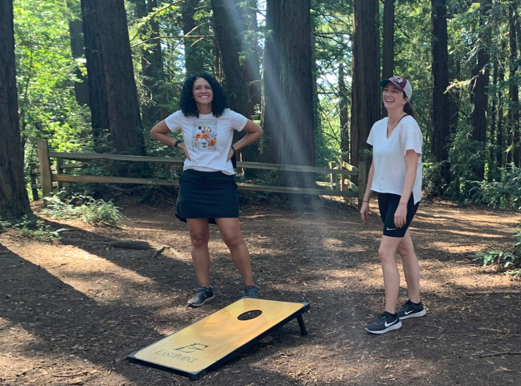 Dana Cilono and Megan De Trane pictured smiling while playing cornhole with redwood trees in the background.