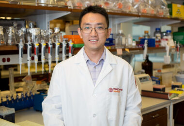 Dr. Chun-Jun Guo smiling in front of his lab with a row of pipettes behind him.