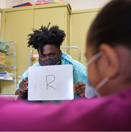 Tutor holds a whiteboard with an "R" written on it