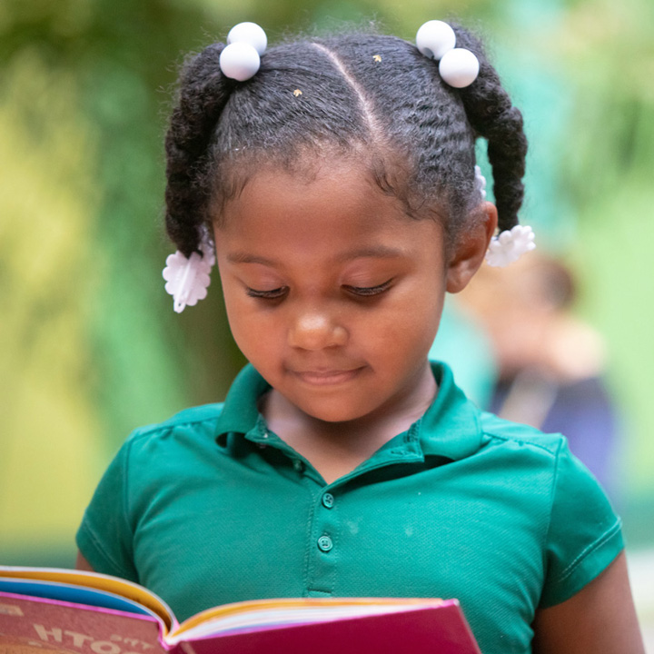 A young Black girl smiling while she looks down at an open book, reading.
