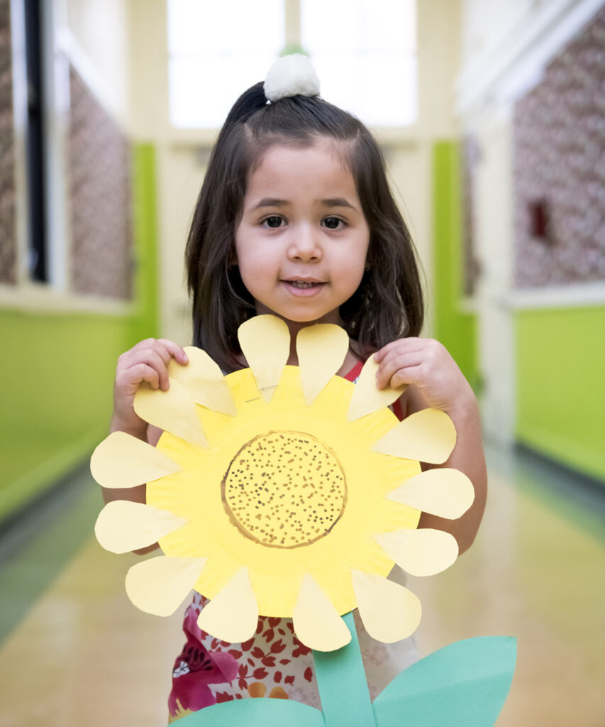 A young girl smiling and holding up a large sunflower she made out of colored paper.