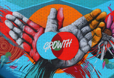 A bright and colorfully painted mural a building shows two overlapping hands with the word growth in an intersecting venn diagram on the palms.