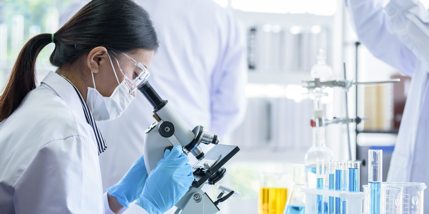 An Asian woman wearing a white lab coat, blue latex gloves, a face mask and goggles looking into a microscope.