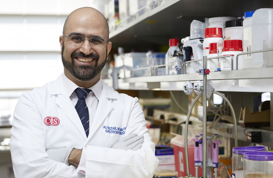 Dr. Ali Rezaie smiling in his lab, wearing a white lab coat, with lab equipment in the background.