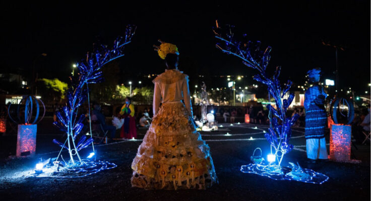 Person in an elaborately designed floor-length dress and hat stands under a sculpture of two branches curving towards center, illuminated by blue light.