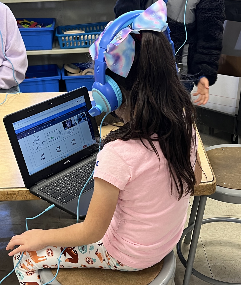 A student wearing headphones sits with a laptop and fills in the missing letter on a series of three-letter words (cat, tag, cap)
