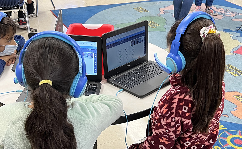 Two young students wear bright blue headphones, while look at an interactive lesson on a laptop.