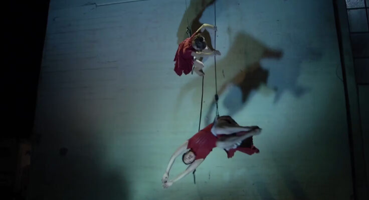 Two aerial dancers wearing red and hanging from a rope on the side of a white building.