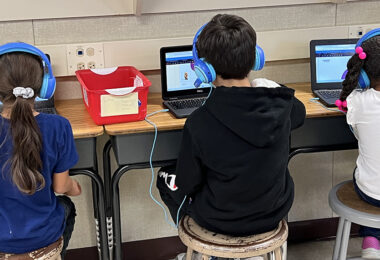Three students seated at adjacent desks wear bright blue headphones and engage with their laptops. Their backs face toward the camera.