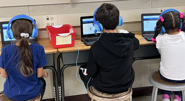 Three students seated at adjacent desks wear bright blue headphones and engage with their laptops. Their backs face toward the camera.